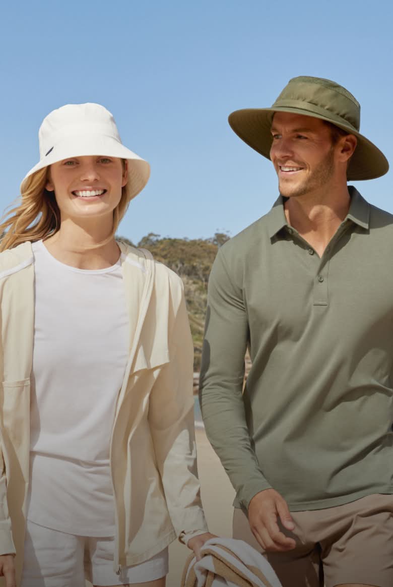 A woman and a man walking along a beach on a sunny day wearing sun hats.