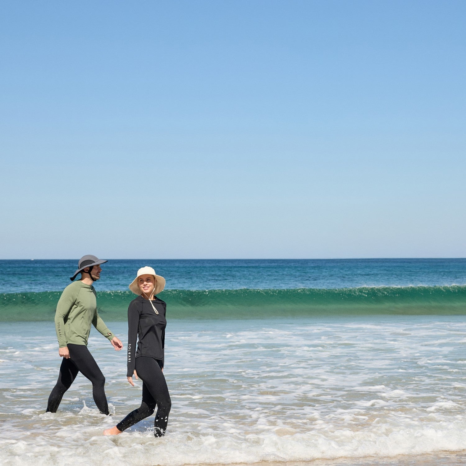 A couple walking along the sea edge in Solbari clothing and sun hats.