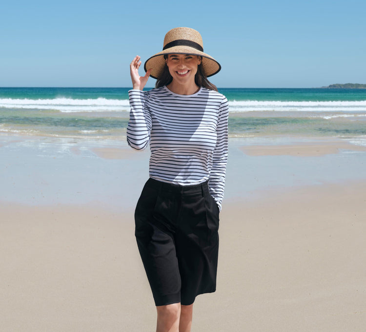 A woman on a beach wearing a Solbari sun protective UPF 50+ raffia sun hat and long sleeve t-shirt, with black shorts.