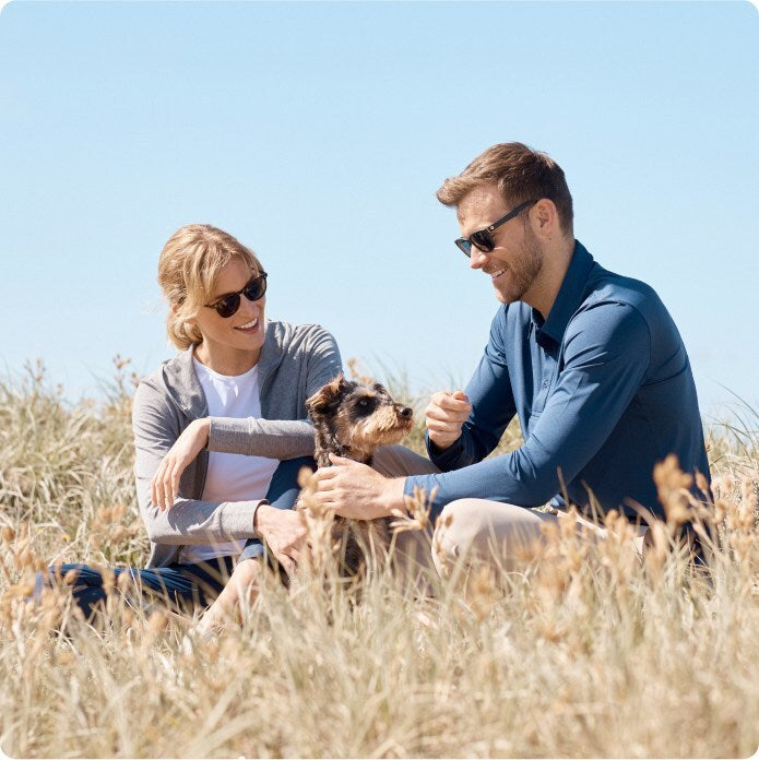 A woman and a moan sitting in the grass with a dog, with blue sky behind them.