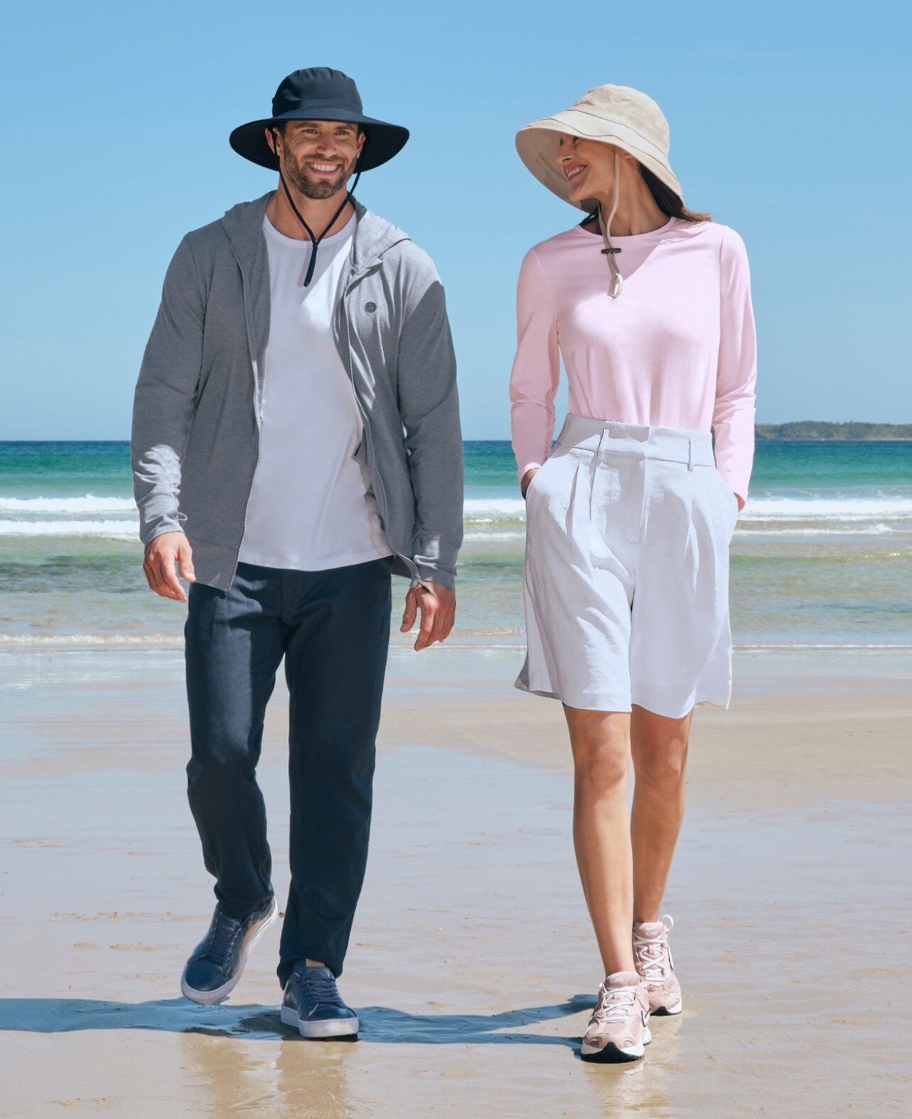 A man and a woman walking on the beach wearing Solbari UPF 50+ sun hats and sun protective clothing.